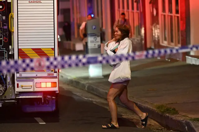 Seorang perempuan membawa bayinya di kawasan Bondi Beach usai penembakan terjadi, dengan garis polisi yang sudah dibentangkan di lokasi kejadian.
