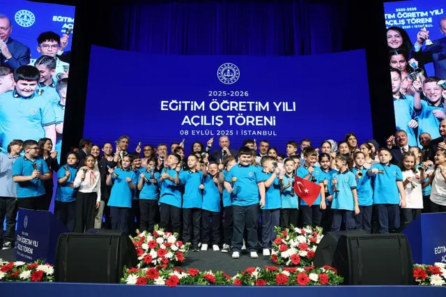 Cumhurbaşkanı Erdoğan mavi bir perdenin önünde mikrofona konuşuyor, önünde çok sayıda çocuk var.