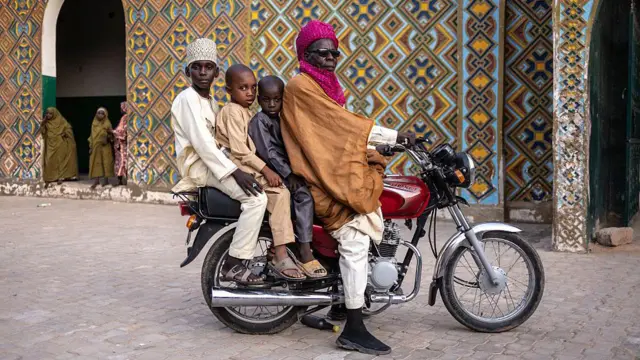 A man and three children sit on a motorbike. An intricately-patterened building can be seen behind them.
