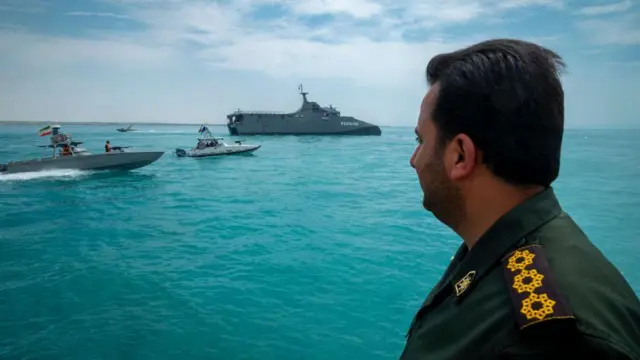 Devrim Muhafızları Ordusu'na bağlı Deniz Kuvvetleri'ne bağlı bir rütbeli asker, geçit törenini takip ediyor arkada, deniz araçları var.