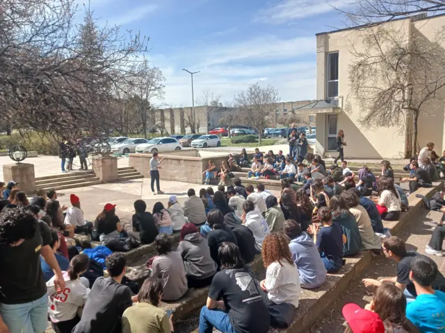 Bir grup öğrenci üniversite bahçesinde bir kişiyi dinliyor.