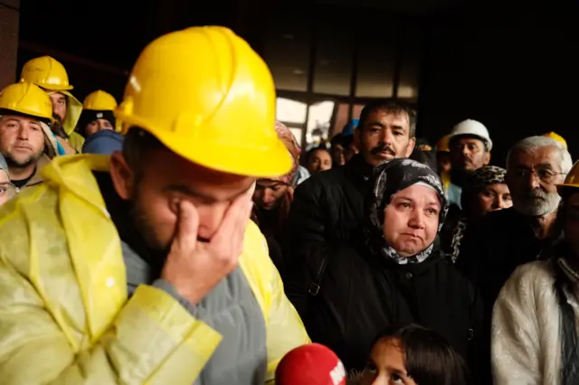 Sarı baret ve yağmurluk giymiş bir madenci gazetecilerin sorunlarını yanıtlarken ağlıyor. 