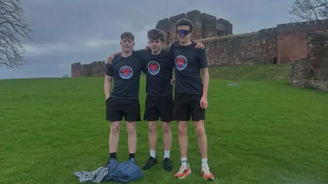 Theo, Oliver y Jacob están de pie frente al Castillo de Carlisle, abrazados. Es un día gris y están de pie sobre césped verde. Todos llevan pantalones cortos negros y camisetas azul oscuro con un símbolo circular y un corazón rojo dentro. Todos tienen el pelo castaño oscuro. Jacob lleva gafas de sol.