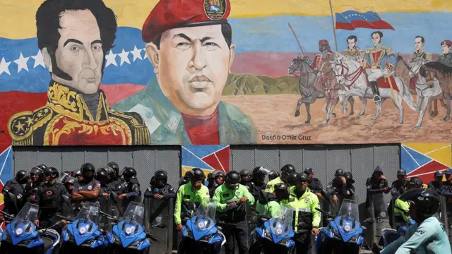 Hugo Chavez'in bere taktığı devasa bir duvar resminin önünde, kask takmış polis memurları bisikletlerin arkasında sıralanmış duruyorlar.