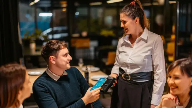 Клієнт оплачує рахунок у ресторані, торкаючись телефоном платіжного пристрою, який тримає офіціантка