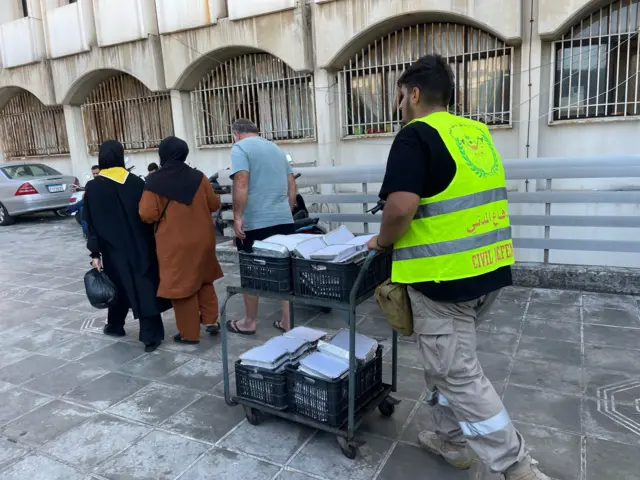 Um jovem com um coletealta visibilidade empurra um carrinho cheiorefeições, enquanto àfrente dois idosos seguram o braço um do outro para caminhar.