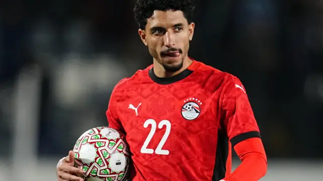 Omar Marmoush, visible de la taille vers le haut, vêtu d'un maillot rouge égyptien avec un numéro 22 blanc sur la poitrine, met sa langue sur le côté gauche de sa bouche alors qu'il tient un ballon de football entre sa main droite et son corps