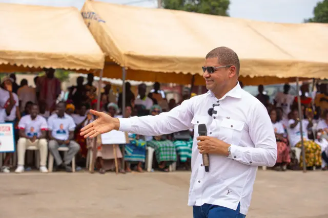 Jean Louis Billon en chemise blanche et lunettes e soleil tient un micro. A l'arrière-plan on voit une rangée de personnes assise portant des t-shirts à son effigie