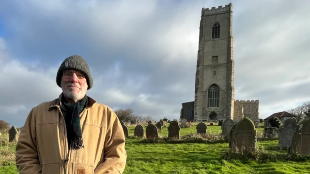 Un hombre con chaqueta color arena, gorro de lana gris y bufanda oscura mira a la cámara en un cementerio. Tiene barba gris y gafas. Detrás de él se ven una iglesia medieval gris, una torre y lápidas. El cielo está nublado.