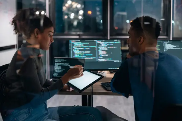 Una mujer y un hombre están trabajando en una habitación donde hay varias pantallas de computador con sistemas de IA. 