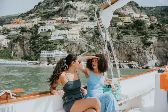 Dos mujeres visiblemente relajadas y felices, disfrutan de un paseo en barco por el mar Mediterráneo. Al fondo se divisa la famosa vista de Positano, Italia.