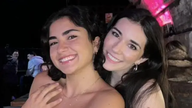 Duas garotas sorrindo para a câmera. À esquerda, a garota tem cabelo escuro na altura dos ombros. Ela está usando uma blusa jeans azul e um colar prateado. A garota à direita tem cabelo escuro e longo. Ela está usando uma blusa jeans azul e brincos prateados. A foto foi tiradaque signifie cbetum bar com longas mesasque signifie cbetmadeira ao fundo.