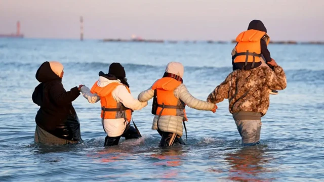 семья - родители и трое детей в спасательных жилетах - заходят в воду, вероятно, направляясь к лодке, которая за кадром