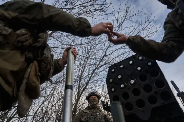 Військові працюють із пусковою системою. Один з них тримає довгу металеву жердину чи стрижень, а інший простягає руки, ніби передає дрібний предмет або допомагає закріпити щось на цьому обладнанні. Праворуч видно багатозарядний блок установки із круглими отворами для боєприпасів. Позаду стоїть ще один військовий у шоломі та повному спорядженні. У тлі — голі гілки дерев і відкрите небо.