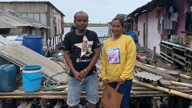 Elik dan Apriani berdiri di depan rumah mereka yang tengah dibangun kembali setelah roboh terseret arus.