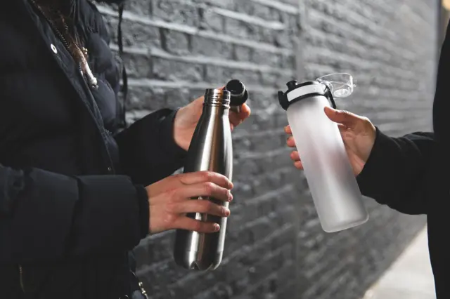 Dua orang sedang menggenggam dua jenis botol yang berbeda.