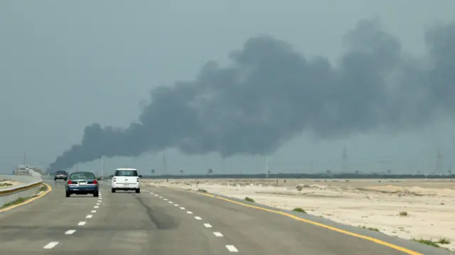 Three cars drive along a three-lane highway with smoke ahead of them. The sky is dark blue and the sides of the road are dusty with a few shrubs.