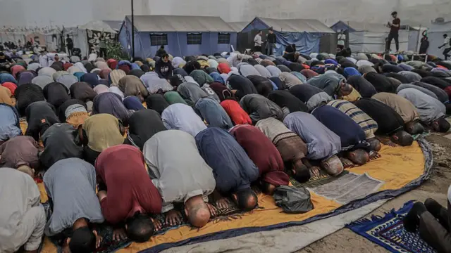 A view of a group of praying men bending to pray in muslim way wit different colours of clothes. Makeshift tent dey show for di back.