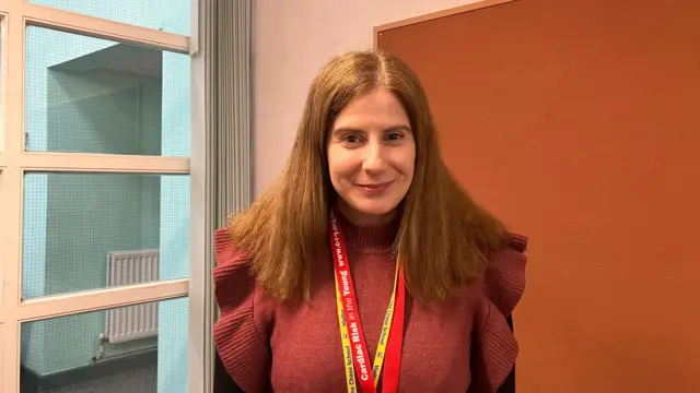 Se muestra una mujer con cabello largo y rojo que viste un suéter rojo con volantes con un tablero de anuncios marrón detrás de ella y una ventana a su lado derecho.