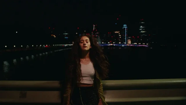 دیبا حکمت در نقش زیبا در صحنه‌ای از فیلم در شب به حصاری تکیه داده، چشم‌هایش بسته است و چشم‌انداز پشت سر او ساختمان‌ها با چراغ‌های روشن است