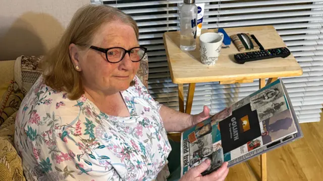 Uma mulher de óculos, com cabelos castanho-claros já grisalhos, segura um álbum de recortes com fotos do filho coladas na capa