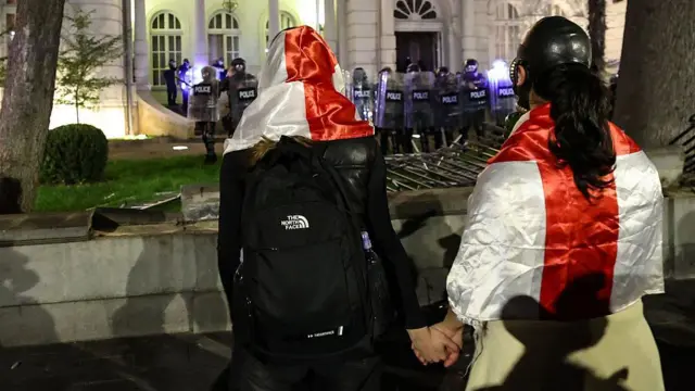 Демонстранты держатся за руки, стоя напротив сотрудников спецназа у президенского дворца в Тбилиси. 4 октября 2025 года 