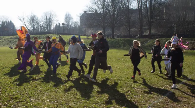 La gente corre por un campo sosteniendo sartenes.