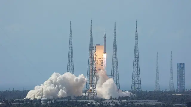 A dramatic launch photograph taken from ground level shows a large white rocket rising from its launch pad on a column of brilliant orange and white fire. Dense clouds of white smoke and steam billow outward from the base of the rocket across the concrete launch platform, which sits within a tropical coastal landscape — palm trees and a clear blue sky are visible in the background