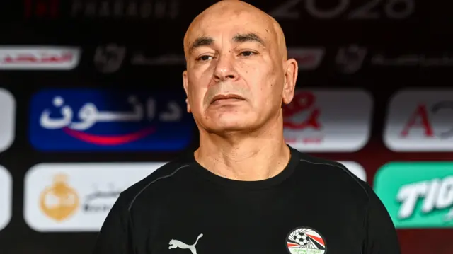 Hossam Hassan vu de la poitrine vers le haut portant un haut d'entraînement noir égyptien alors qu'il est vu devant un abri affichant divers sponsors. 