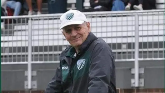 Питер Мигер опирается на низкий белый забор. Он одет в серую куртку с зеленым логотипом регби и белую солнечную шляпу с тем же логотипом. За его спиной зрители сидят на белых трибунах.