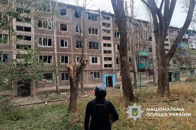 поліцейський стоїть спиною до камери на фоні зруйнованої обстрілами багатоповерхівки 