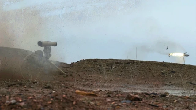 یک نیروی نظامی در حال شلیک موشک ضدتانک کورنت است، موشک با شعلهای از پشت سرش، در فاصله کم از پرتابگر شلیک شده، و سرباز سر خود را پایین گرفته است دود و غبار ناشی از شلیک دیده میشود