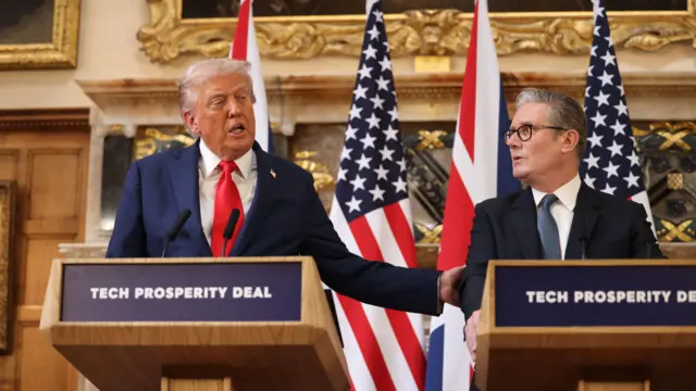 Трамп за деревянной трибуной в белой рубашке, красном галстуке и синем пиджаке держит за руку Стармера, стоящего за соседней трибуной в черном пиджаке, белой рубашке и синем галстуке. Они стоят на фоне флагов США и Великобритании