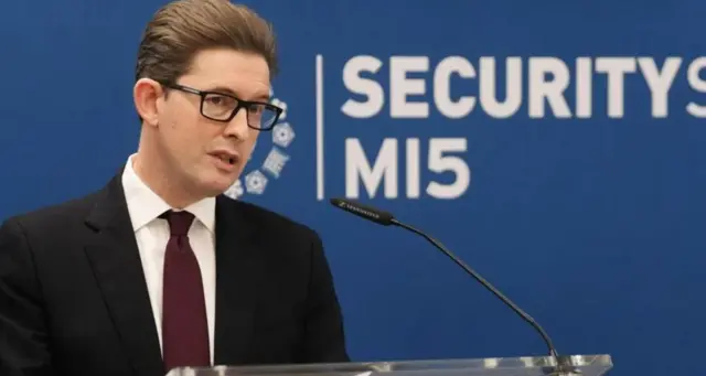 Кен МакКаллам у чорному костюмі на фоні синього банеру 