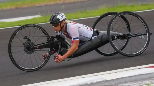 A foto mostra uma mulherquantos saques posso fazer na greenbetspele clara, com um capacete pretoquantos saques posso fazer na greenbetsuma cadeira adaptada para o cilismo Paralímpico. Ela pedala com os braços.