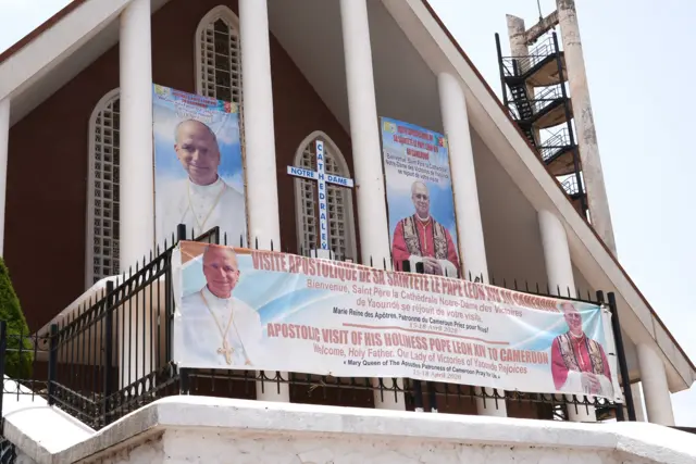 Exterior of Notre Dame Cathedral in Yaoundé wit three posters of Pope Leo XIV dey on display on di building and fence, e dey announce im visit to Cameroon in French.
