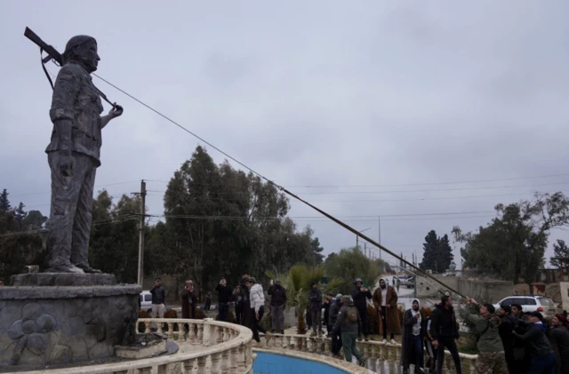 Onlarca erkek kent meydanındaki bir silahlı kadın heykelini yıkmak için hazırlanıyor. Bir halat heykelin boynuna bağlanmış ve erkekler onu çekmeye çalışıyor.