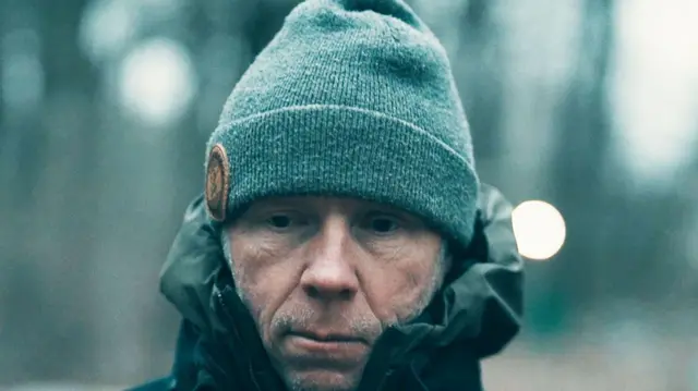 ਗ੍ਰੇਗ ਸਕੁਇਰ ਜੰਗਲ ਵਿੱਚ ਸੈਰ ਕਰ ਰਿਹਾ ਹੈ - ਉਸਨੇ ਇੱਕ ਗੂੜ੍ਹੇ ਰੰਗ ਦਾ ਕੋਟ ਅਤੇ ਸਲੇਟੀ ਰੰਗ ਦੀ ਬੀਨੀ ਟੋਪੀ ਪਾਈ ਹੋਈ ਹੈ