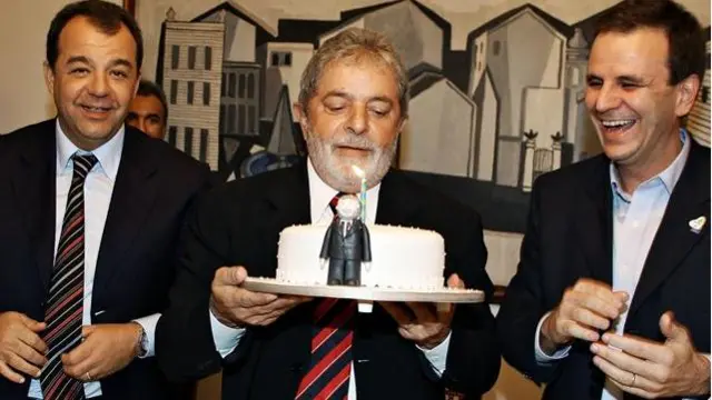 Lula, segurando um bolo de aniversário, entre Sergio Cabral e Eduardo Paes