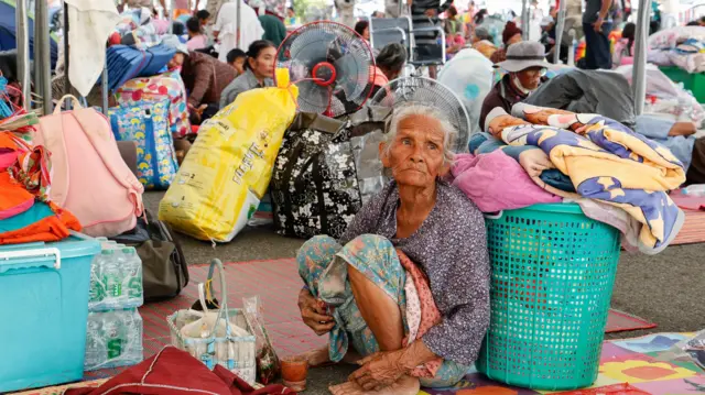 Starsza kobieta o zaniepokojonym wyrazie twarzy siedzi na podłodze obok koszyków, toreb i wachlarzy. W tle widać wiele innych osób z bagażami.