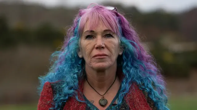 Foto de Jayne, com cabelo comprido pintado de roxo e azul, olhando diretamente para a câmera, com paisagem desfocada de uma montanha ao fundo