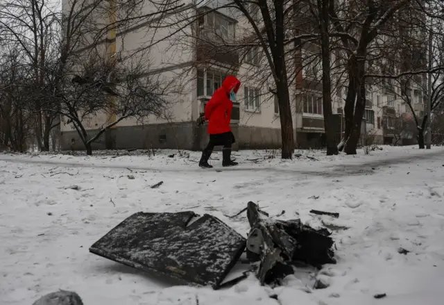 Жінка у червоній куртці йде повз уламок дрона, що лежить на снігу