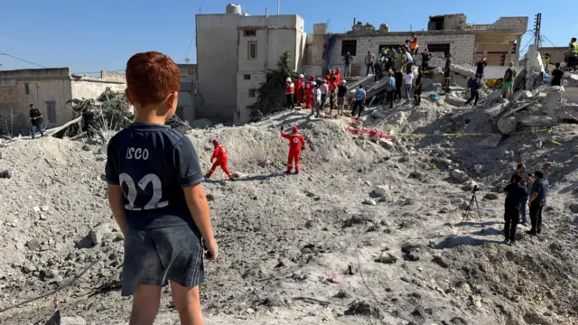 Fotografia colorida mostra criança ruiva de costas, com uma camiseta de time de futebol, olhando os escombros de local atingido por bombas no Líbano. Moradores, funcionários e jornalistas são vistos no transitando no fundo da imagem.
