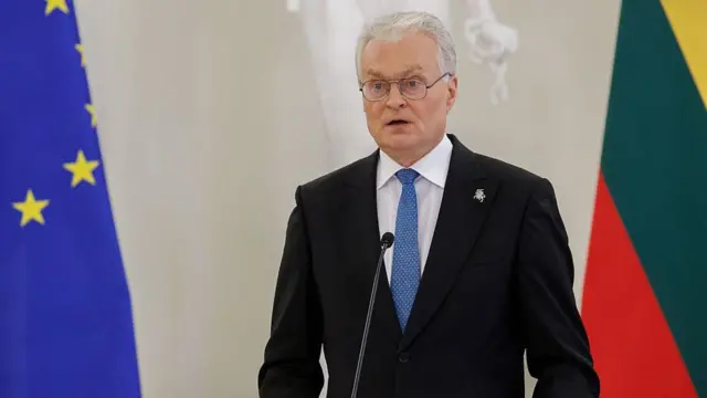 Президент Литвы Гитанас Науседа за трибуной на фоне флагов Литвы и Евросоюза. Он носит очки и гладко выбрит, волосы седые. На нем черный пиджак и белая рубашка с синим галстуком.