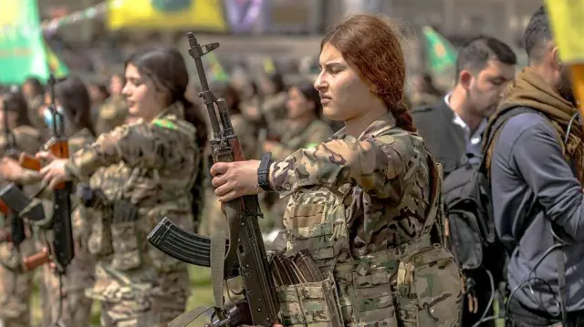 Kamuflajlı kadın askerler ellerinde AK-47 tüfekleriyle tören duruşunda. Arkada sarı ve yeşil renki bayraklar görünüyor. Kadınların başları açık ve saçları örgülü.
