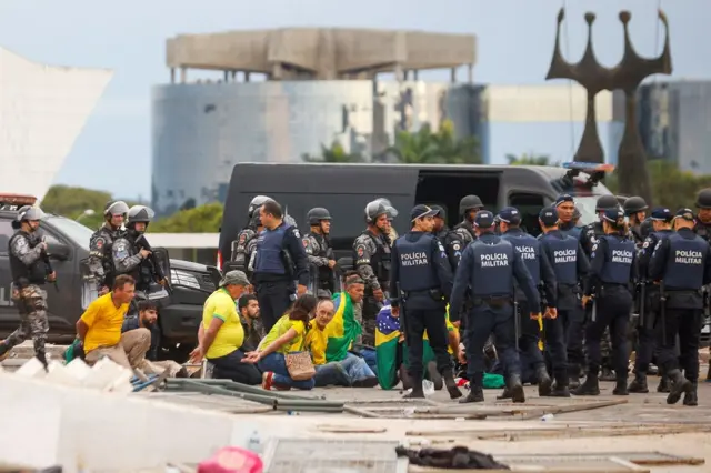 Pessoas de camisa verde e amarela sentadas com as mãos algemadas; eles estão rodeados por policiais na Praça dos Três Poderes