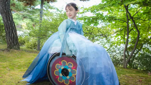 یوه کوریایۍ ښځه په یوه زرغون ځنګل کې پر څوکۍ د واده په دودیزو کالیو هانبوک کې ناسته ده او ویلچېیر ټایرونه هم په ګلانو رنګ شوي دي.