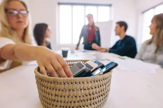 Um grupo de pessoas reunidas em torno de uma mesa sobre a qual há uma cesta com vários celulares