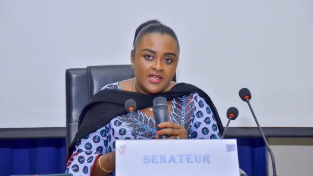 Francine Muyumba yicaye arimo kuvugira mu ndangururamajwi nini, imbere ye hari izindi ndangururamajwi eshatu ntoya. Yambaye ikanzu yo mu gitenge, yiteye fulari y'umukara ku ijosi imbere no ku ntugu, afite umusatsi ubinditse ufungiye inyuma.
