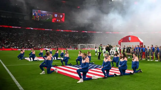 jugadoras de las selecciones de EE.UU. e Inglaterra se arrodillan junto a las banderas de sus países extendidas en el suelo de un estadio